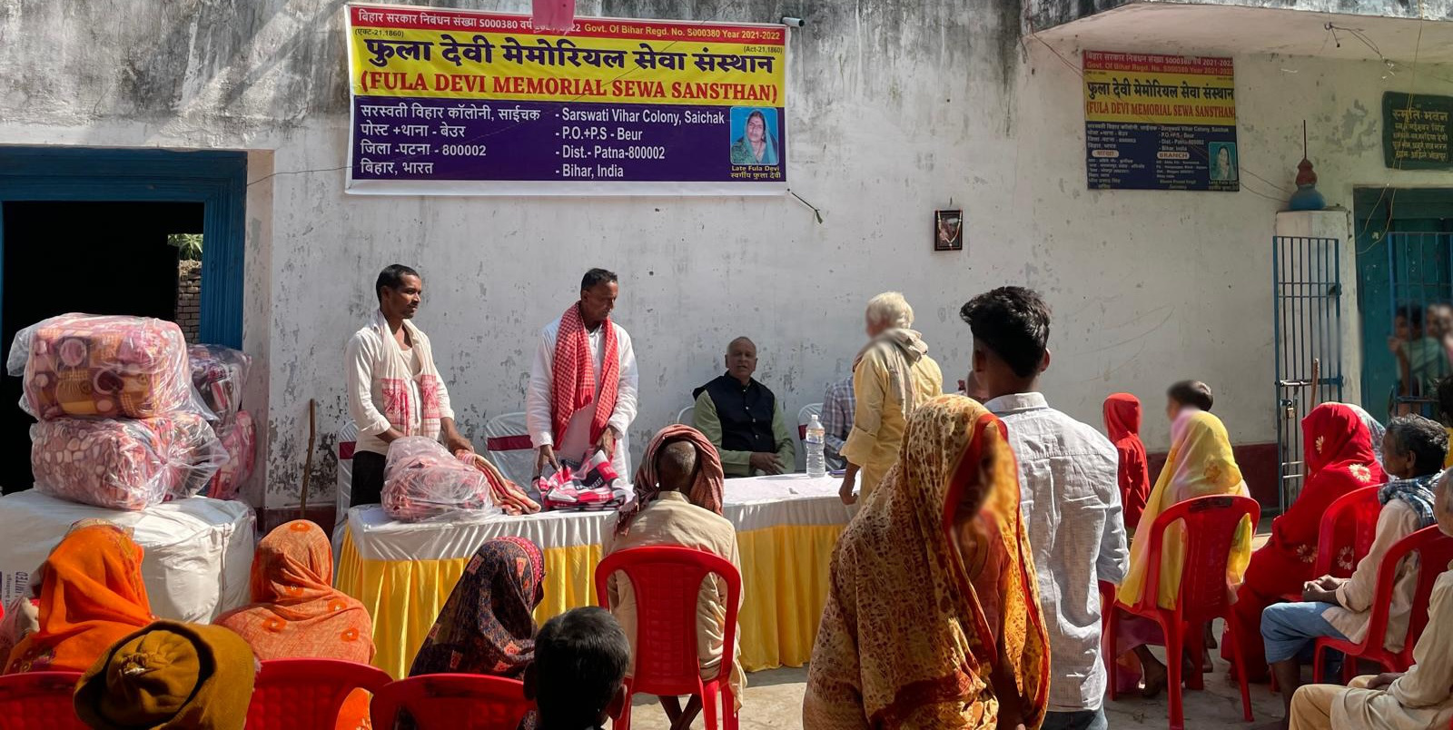 Blankets stacked beside a table where members of the institution hand out items to residents seated in a courtyard, under a banner bearing the institution's name.